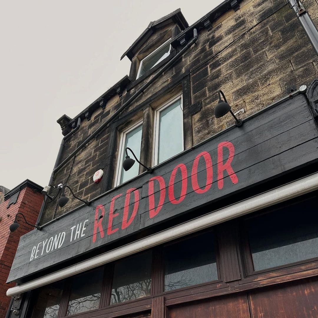 Beyond The Red Door historic building tourism attraction red door signage Signage of "Beyond The Red Door" on a stone building, featuring large red letters against a dark wooden background. The building has multiple windows and is set against a gray sky.