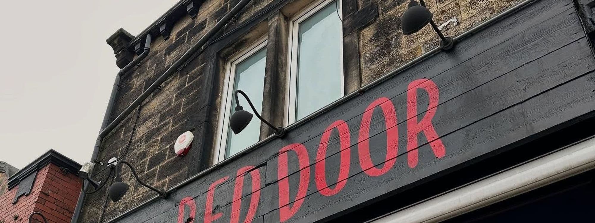 Alt text: Exterior view of the "Beyond The Red Door" building, featuring a prominent red sign and windows against a stone wall.