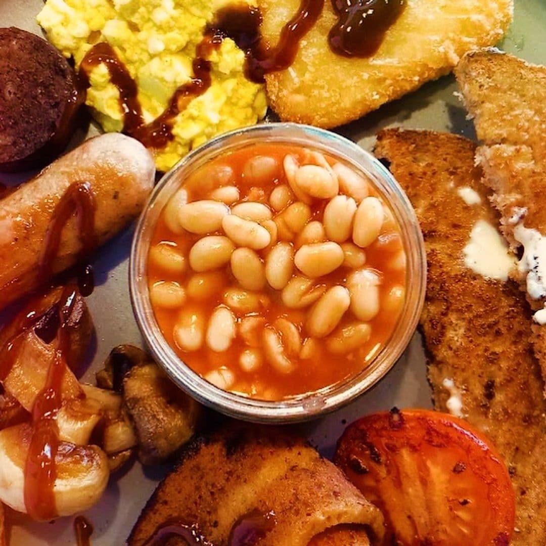 Scoffs Feel Good Food breakfast platter delicious savoring comfort food A delicious plate of breakfast items from Scoffs Feel Good Food, featuring baked beans in a small glass bowl, scrambled eggs, a hash brown, grilled mushrooms, sausage, and crispy bacon, all drizzled with sauce.