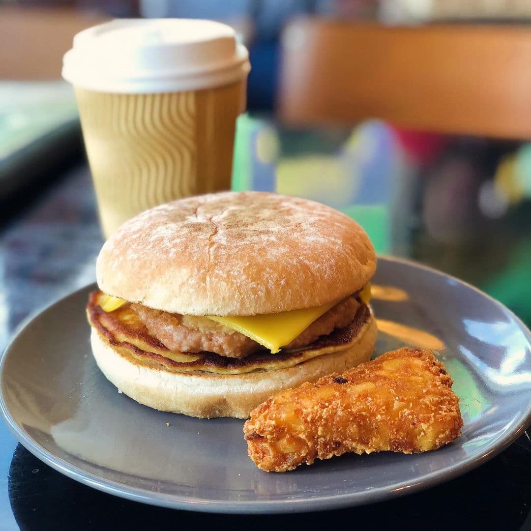 Scoffs Feel Good Food breakfast sandwich coffee snack A delicious breakfast sandwich featuring a sausage patty and melted cheese on a toasted bun, served with a crispy hash brown and a cup of coffee. Scoffs Feel Good Food.