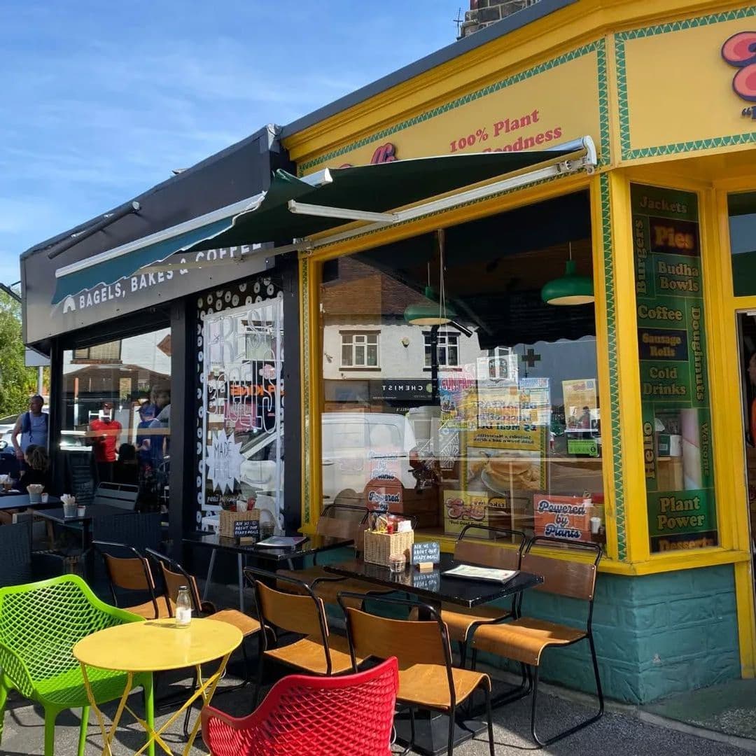 Scoffs Feel Good Food cafe outdoor seating colorful exterior **Scoffs Feel Good Food**: A vibrant café exterior featuring colorful outdoor seating with green, yellow, and red chairs. The café showcases a large window displaying a menu with various food options, including bagels and plant-based dishes. The bright yellow and green awnings add to the cheerful atmosphere, inviting visitors to enjoy a meal outside.