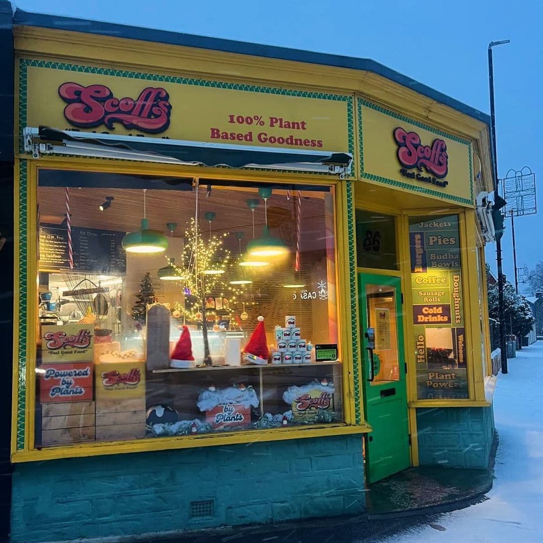 Scoffs Feel Good Food plant based cafe winter snowy exterior Alt text: Exterior view of Scoffs Feel Good Food, a vibrant plant-based eatery featuring a colorful sign and festive decorations in the window. Snow covers the ground, creating a cozy winter atmosphere.