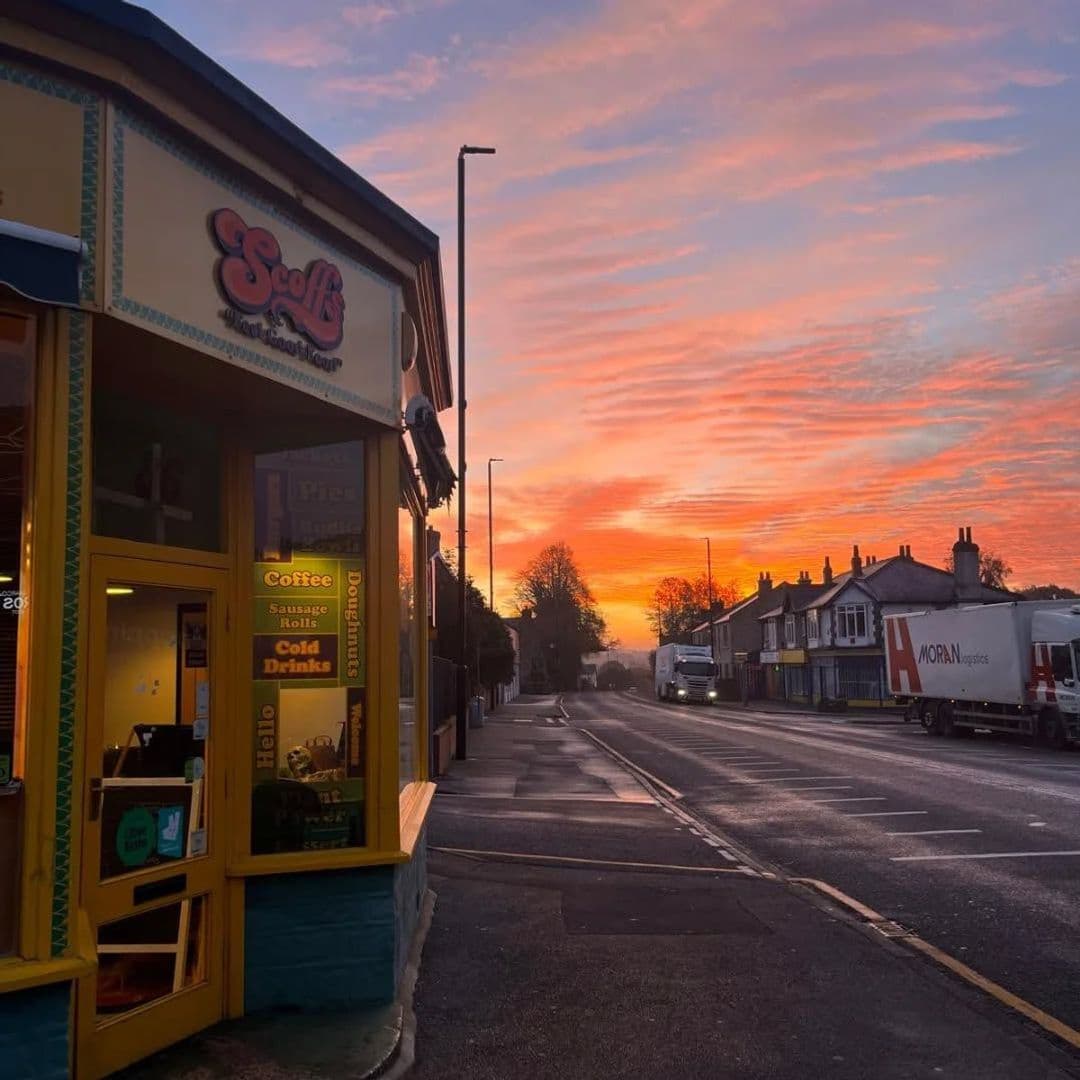 Scoffs Feel Good Food sunset coffee cafe street view Sunrise view outside Scoffs Feel Good Food, showcasing vibrant orange and pink skies. The image features the café's colorful exterior with signage for coffee, sausage rolls, and cold drinks. A quiet street is visible with parked trucks and trees lining the road.