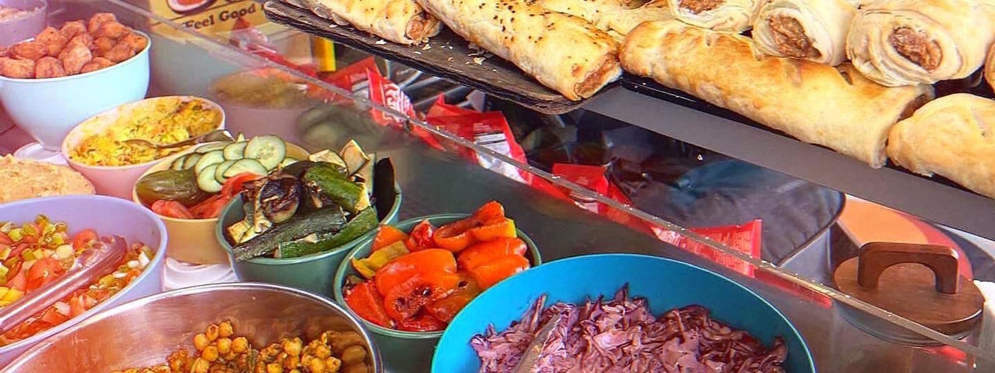 A vibrant display of fresh dishes at Scoffs Feel Good Food, featuring colorful bowls of salads, roasted vegetables, and a variety of snacks. In the background, golden-brown pastries and rolls are neatly arranged, inviting customers to enjoy a delicious meal.