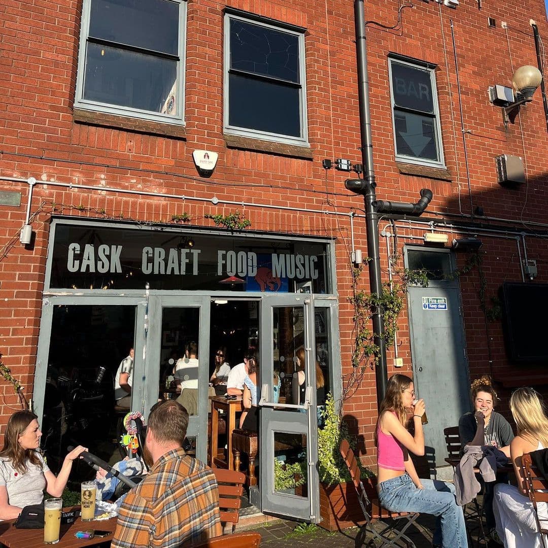 The Head of Steam Headingley cask craft food music outdoor dining The Head of Steam Headingley features a lively outdoor seating area with patrons enjoying drinks and conversation. The entrance showcases the words "CASK CRAFT FOOD MUSIC" prominently on the glass doors, inviting visitors to experience the vibrant atmosphere. The red brick exterior adds a warm touch to the venue, while greenery enhances the inviting ambiance.