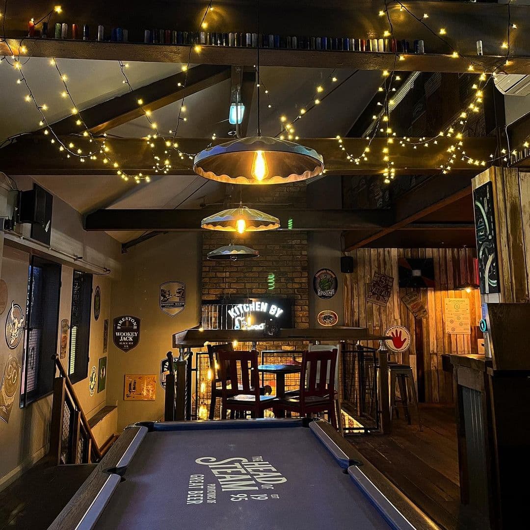 The Head of Steam Headingley cozy ambiance bar interior nightlife Alt text: Interior view of The Head of Steam Headingley, featuring a cozy atmosphere with warm lighting, a pool table in the foreground, and wooden furnishings. The walls are decorated with various beer signs, and a bar area is visible in the background.