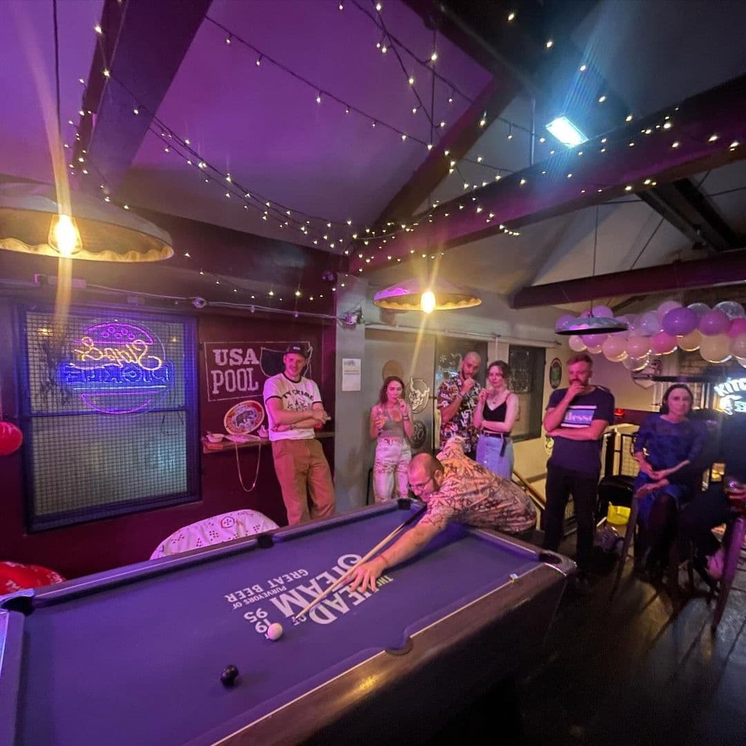 The Head of Steam Headingley pool game night friends fun entertainment A lively scene at The Head of Steam Headingley, featuring a group of people gathered around a pool table. One person is taking a shot, while others watch and engage in conversation. The room is warmly lit with string lights, creating a cozy atmosphere. Decor includes a neon sign and playful decorations, enhancing the inviting vibe of the venue.