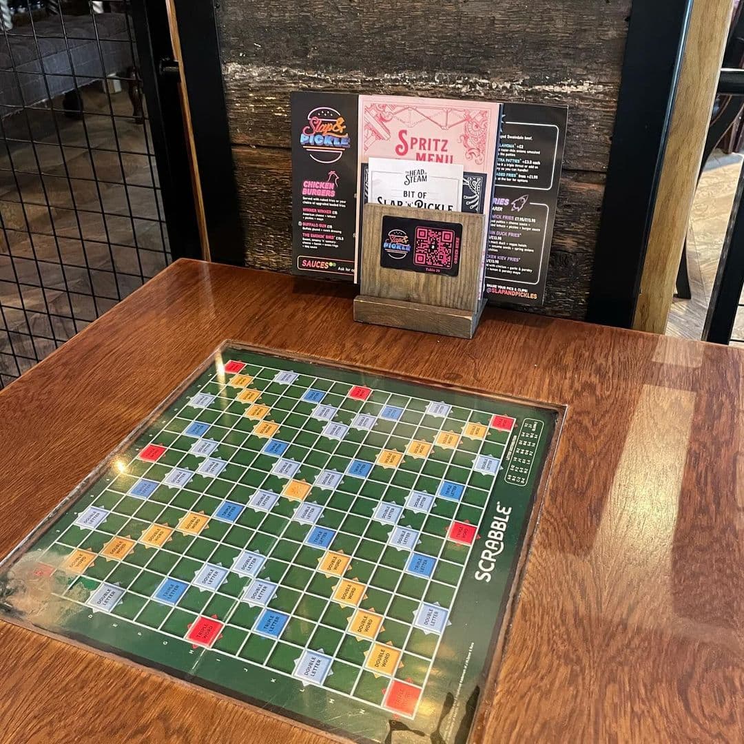The Head of Steam Headingley scrabble game menu relaxation dining experience A wooden table at The Head of Steam Headingley features a Scrabble board under glass. Nearby, a menu stand displays various food and drink options, including a spritz menu. The rustic setting invites guests to enjoy games and refreshments in a cozy atmosphere.