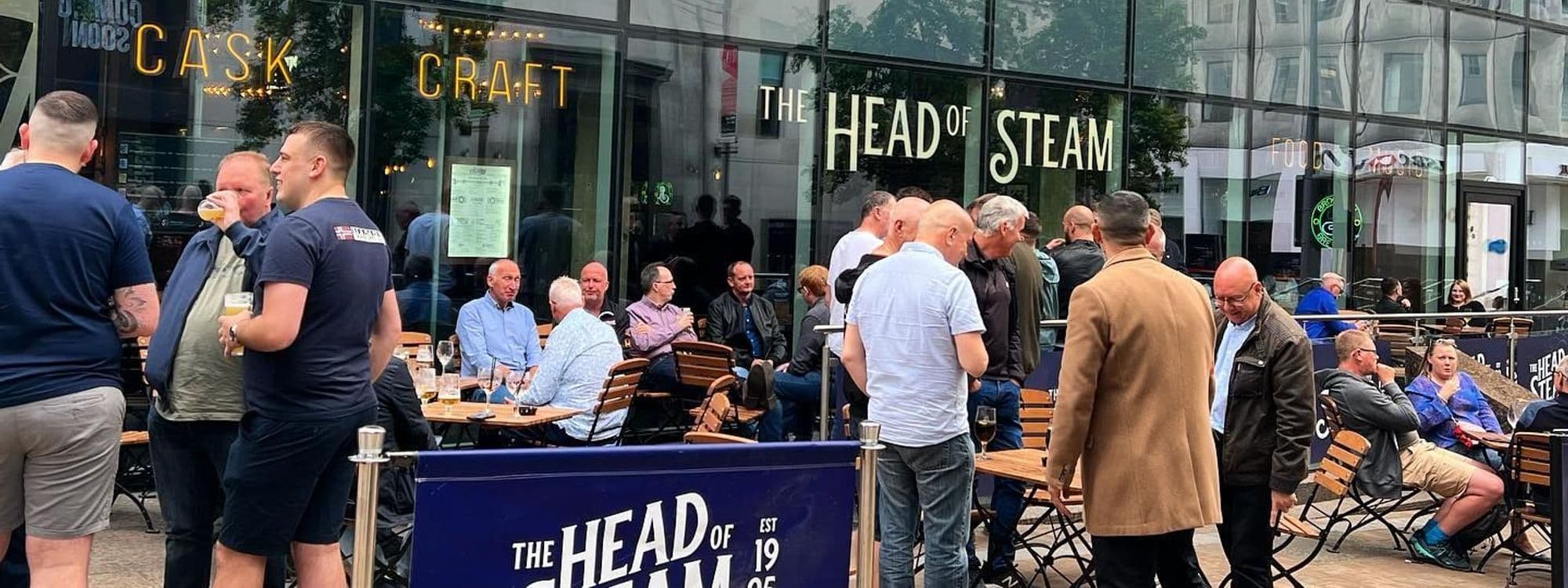 Alt text: A lively outdoor scene at The Head of Steam – Park Row, featuring groups of people socializing and enjoying drinks at tables. The venue's large glass windows display the name "The Head of Steam" and "Craft" in bright lettering, creating a welcoming atmosphere.