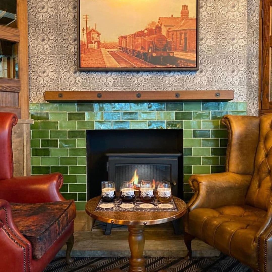 The Head of Steam Park Row cozy fireplace interior drinks relaxation Cozy seating area at The Head of Steam – Park Row, featuring two armchairs, a small wooden table with drinks, and a fireplace with green tiles. A vintage train-themed artwork hangs above the fireplace, adding to the warm atmosphere.