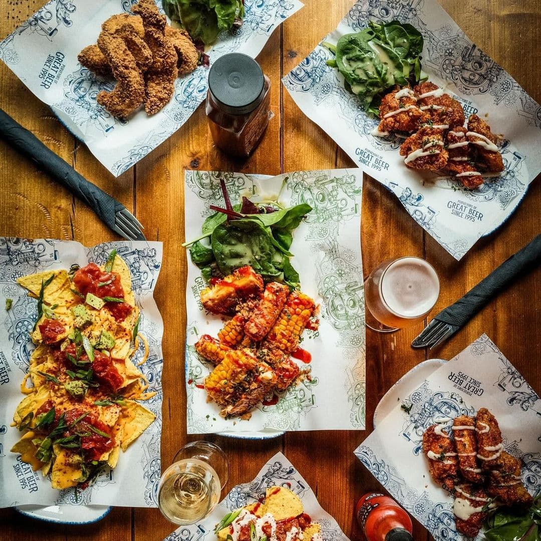 The Head of Steam Park Row delicious foods variety dining experience restaurant Delicious spread of various dishes at The Head of Steam – Park Row, featuring crispy chicken, nachos topped with salsa, grilled corn, and fresh greens. A glass of beer is also visible, set against a warm wooden table.
