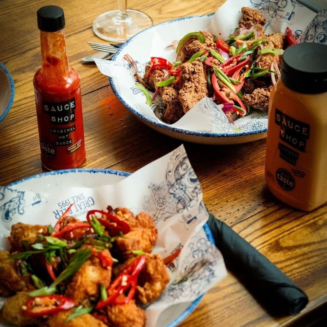 The Head of Steam Park Row fried chicken dishes hot sauce garnish Delicious fried chicken served with fresh vegetables and sauces at The Head of Steam – Park Row. The image features two bowls of chicken, garnished with red chilies and green onions, alongside bottles of hot sauce. A wooden table adds a warm touch to the dining experience.
