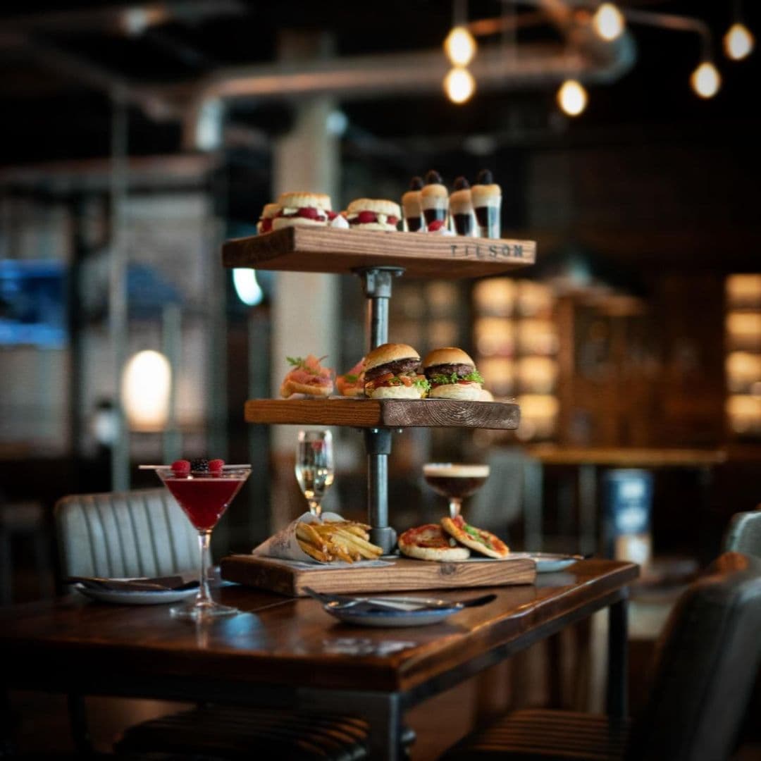 The Head of Steam Park Row gourmet food drinks dining experience modern restaurant Alt text: A beautifully arranged food display at The Head of Steam – Park Row, featuring a tiered wooden stand with gourmet burgers, desserts, and drinks, set on a rustic table in a cozy restaurant atmosphere.