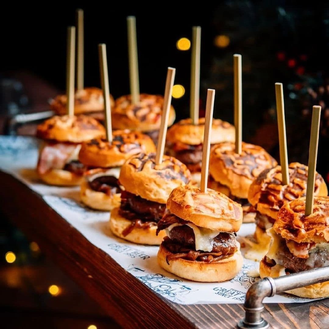 The Head of Steam Park Row gourmet sliders food tasting experience Delicious mini burgers served on a wooden platter with decorative sticks. The burgers feature a variety of toppings and sauces, showcasing a mouthwatering selection. The setting has a warm, inviting atmosphere, perfect for enjoying a meal. This image represents the culinary offerings at The Head of Steam – Park Row.
