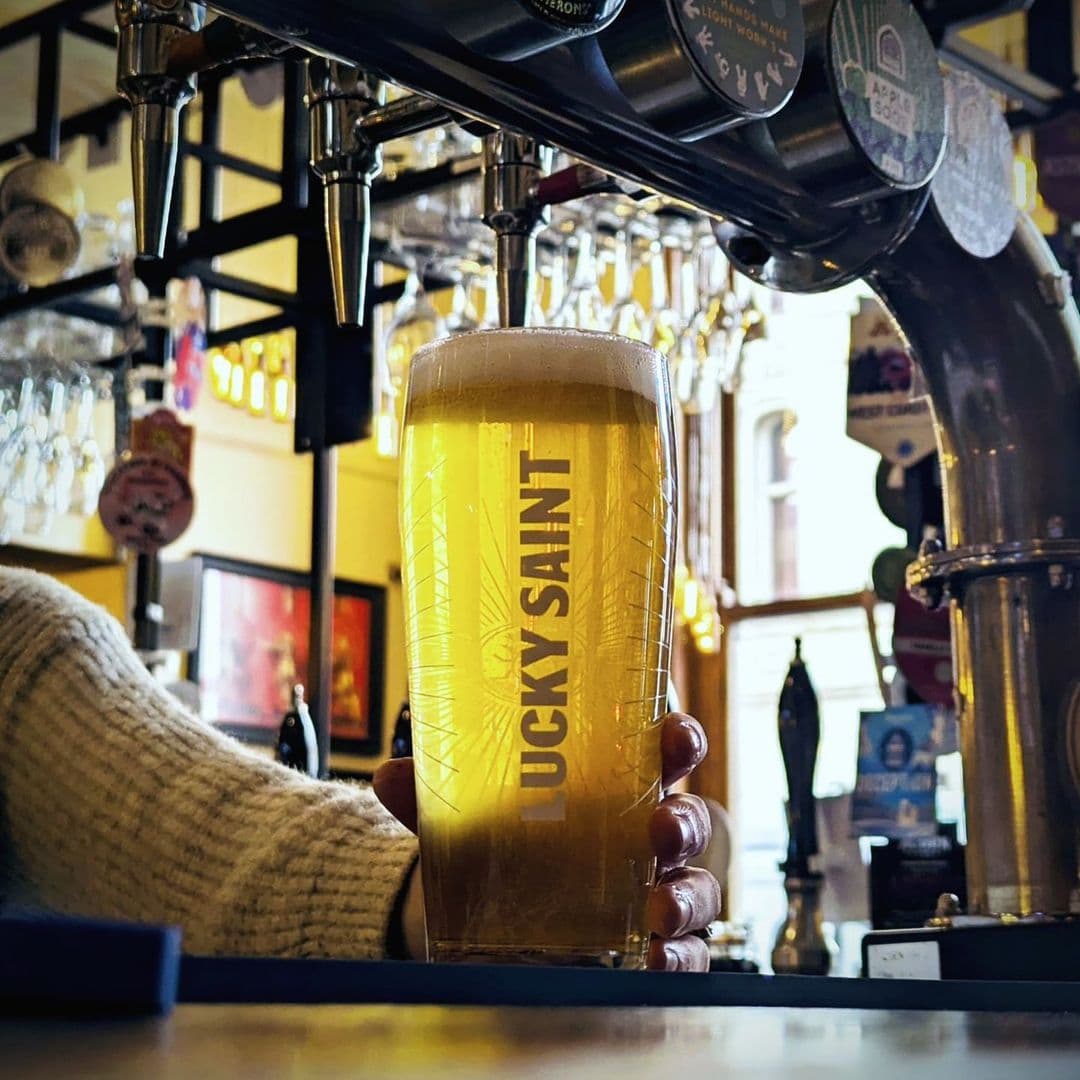 The Head of Steam Park Row lucky saint beer pub experience refreshing drink A hand holding a glass of Lucky Saint beer in a cozy pub setting at The Head of Steam – Park Row. The background features bar taps and shelves filled with glasses, creating a warm and inviting atmosphere.