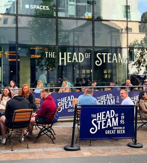 The Head of Steam Park Row outdoor dining great beer social atmosphere urban vibes Outdoor seating area at The Head of Steam – Park Row, with people enjoying food and drinks. The venue features large windows and signage displaying the name and establishment date.