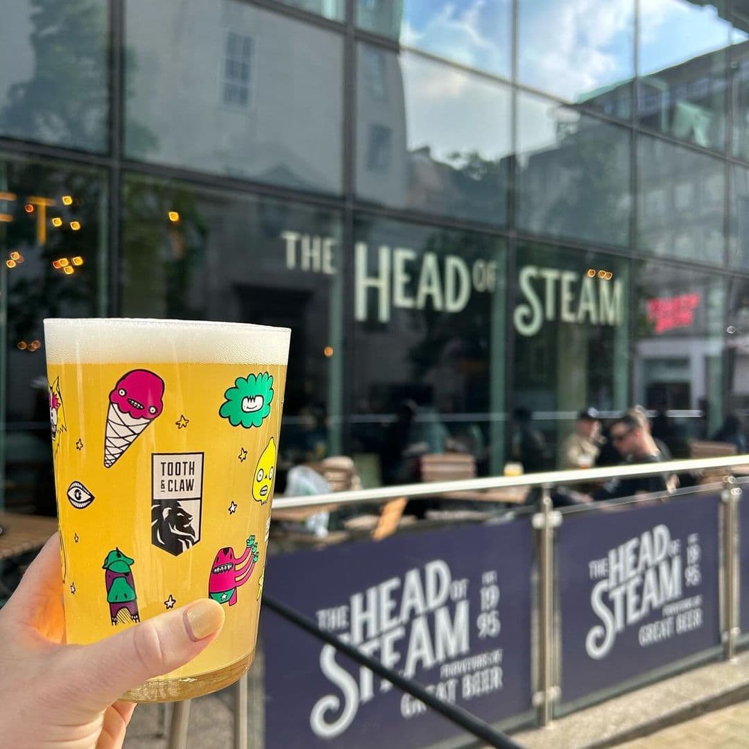 The Head of Steam Park Row vibrant beer outdoor dining relaxation fun A hand holding a colorful pint of beer with fun graphics in front of The Head of Steam – Park Row. The background features the pub's name prominently displayed on a glass facade, with outdoor seating visible.