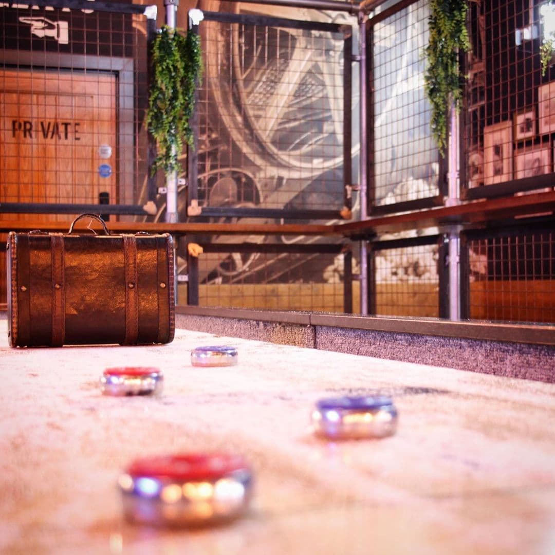 The Head of Steam Park Row vintage suitcase curling game interior **Alt Text:** A vintage suitcase sits on a textured surface with colorful discs scattered around it. The background features a stylish, industrial design with a private area sign and greenery. This scene is located at The Head of Steam – Park Row.