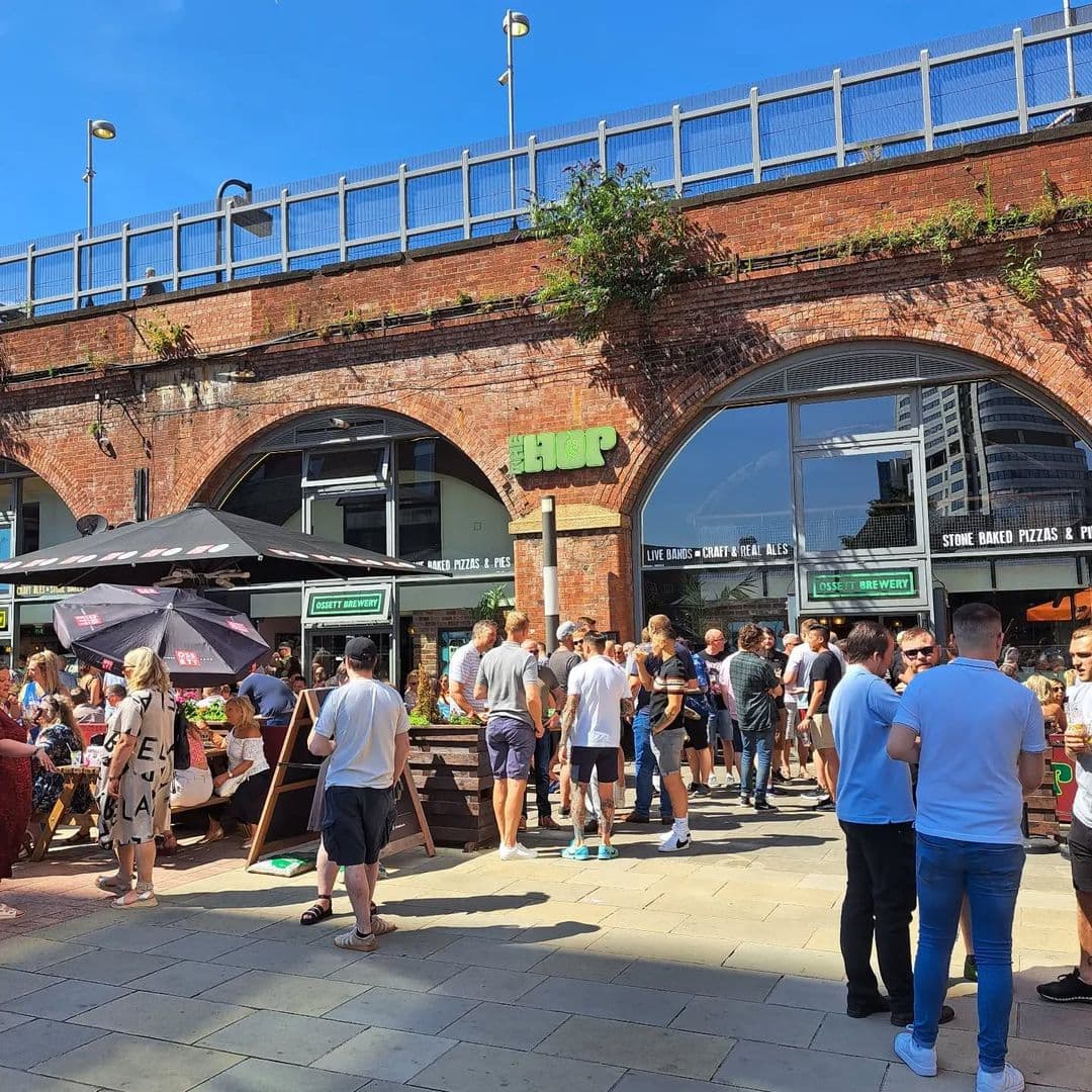 The Hop busy outdoor dining summer festival people A lively outdoor scene at The Hop, featuring a crowd of people enjoying food and drinks. The venue has a rustic brick facade with large windows, and umbrellas providing shade for patrons. The atmosphere is vibrant, with groups socializing and a variety of seating options available. Bright blue skies complete the inviting setting.