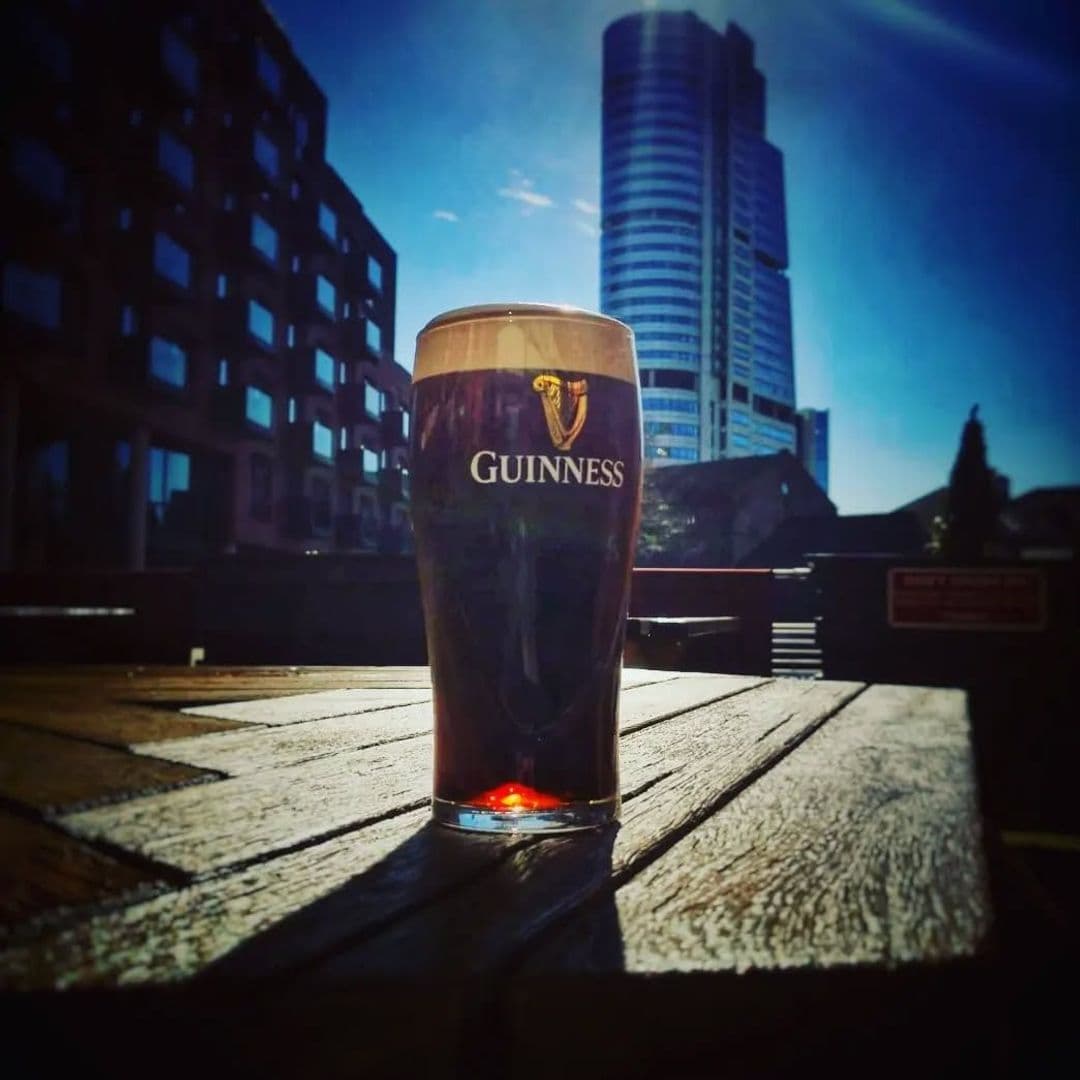 The Hop guinness pint sunny day cityscape A pint of Guinness sits on a wooden table in the foreground, with a bright blue sky and modern buildings in the background. The location is The Hop, a vibrant spot for enjoying drinks and the city atmosphere.