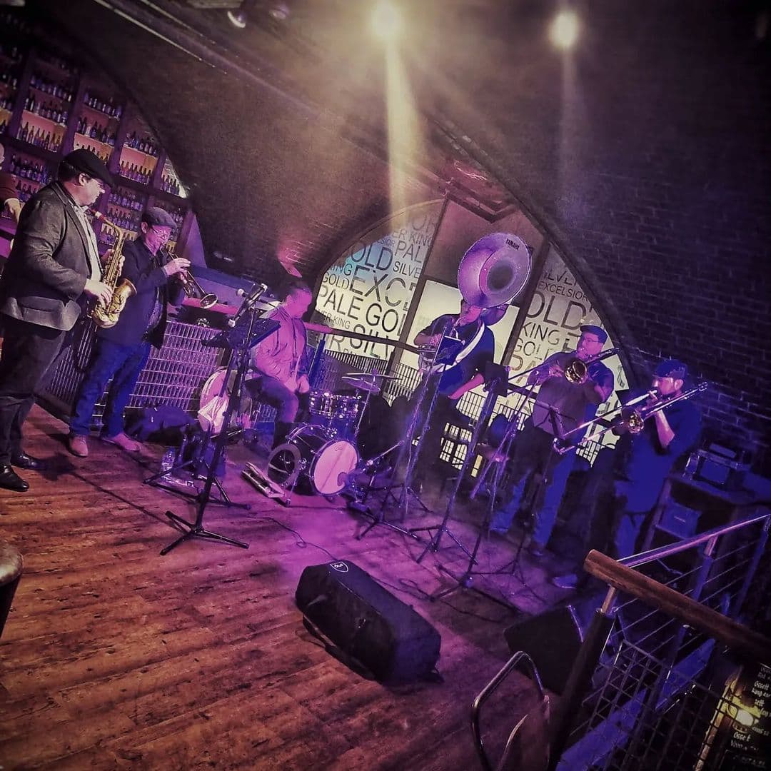 The Hop live music band performance jazz night A lively jazz band performs on stage at The Hop, featuring musicians playing saxophone, trumpet, trombone, and drums. The venue has a warm ambiance with wooden floors and a brick wall backdrop, illuminated by soft lighting.