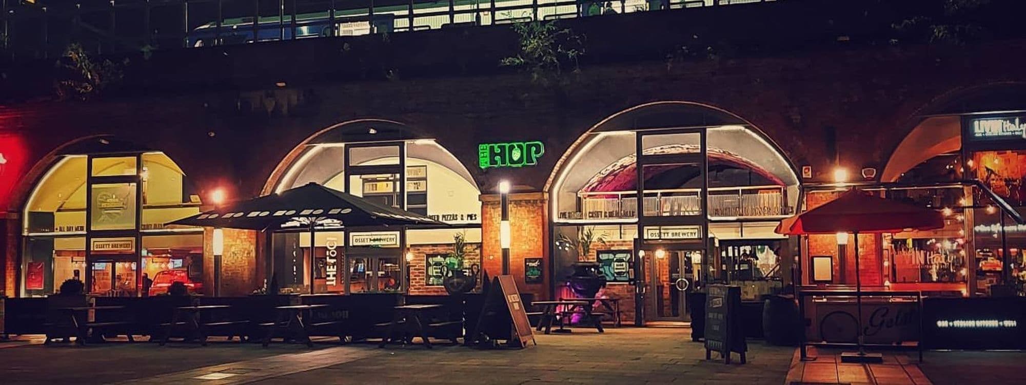 Alt text: The Hop, a vibrant nightlife venue, features illuminated signage and outdoor seating. The building showcases arched windows and brick walls, creating a welcoming atmosphere. It's a popular spot for dining and socializing in the evening.
