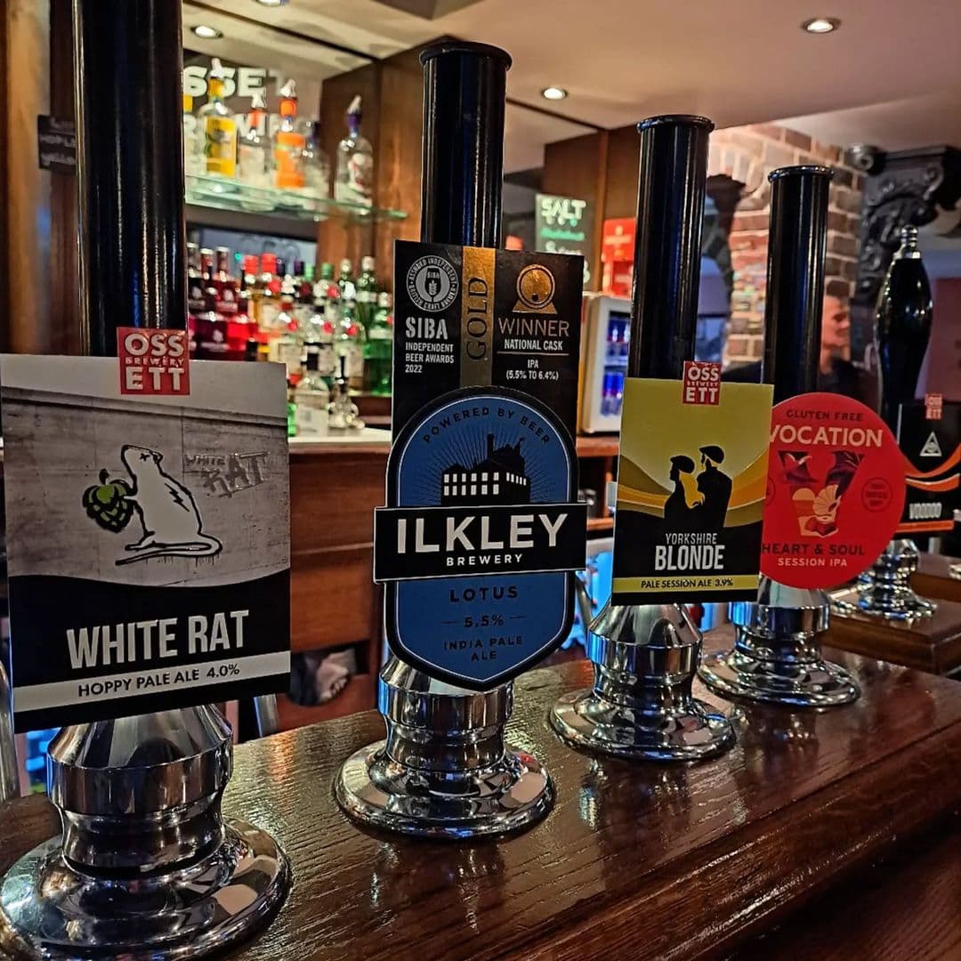 The Hop Alt text: A close-up of beer taps at The Hop, featuring various beer labels including White Rat, Ilkley Brewery Lotus, Yorkshire Blonde, and Vocation Heart & Soul. The background shows a well-stocked bar.