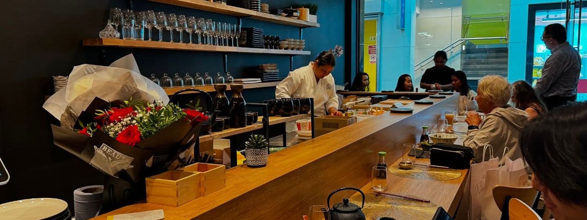 Alt text: A sushi chef prepares dishes at the counter of Fern Modern Sushi. The restaurant features a warm wooden interior, with shelves displaying glassware and plates. Diners enjoy their meals at the counter, surrounded by a cozy atmosphere.