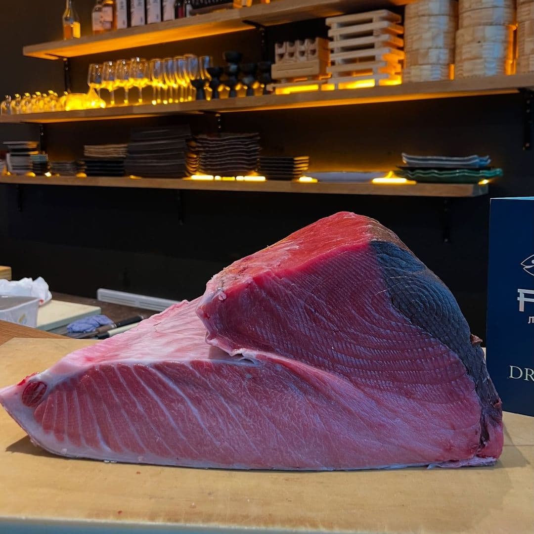 Fern Modern Sushi fresh tuna sashimi preparation japanese cuisine gourmet seafood A large piece of fresh tuna is displayed on a wooden cutting board at Fern Modern Sushi. The tuna has a vibrant pink color with a dark edge, showcasing its quality. In the background, there are shelves with plates and glasses, adding to the restaurant's ambiance.