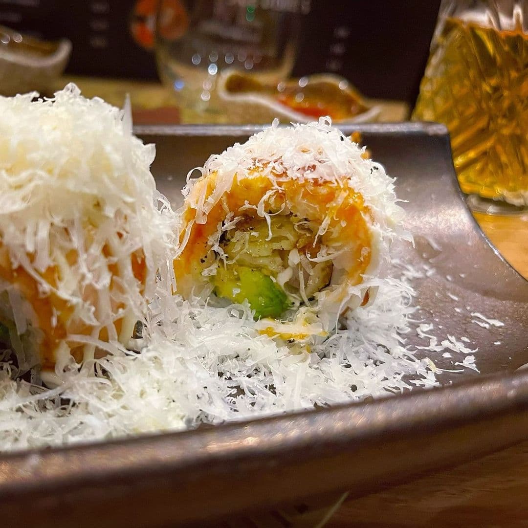 Fern Modern Sushi gourmet sushi rolls fresh ingredients delicious presentation Sushi rolls topped with shredded cheese served on a dark plate at Fern Modern Sushi. The rolls contain ingredients like avocado and shrimp, with a drizzle of sauce. A blurred background shows a warm, inviting atmosphere.