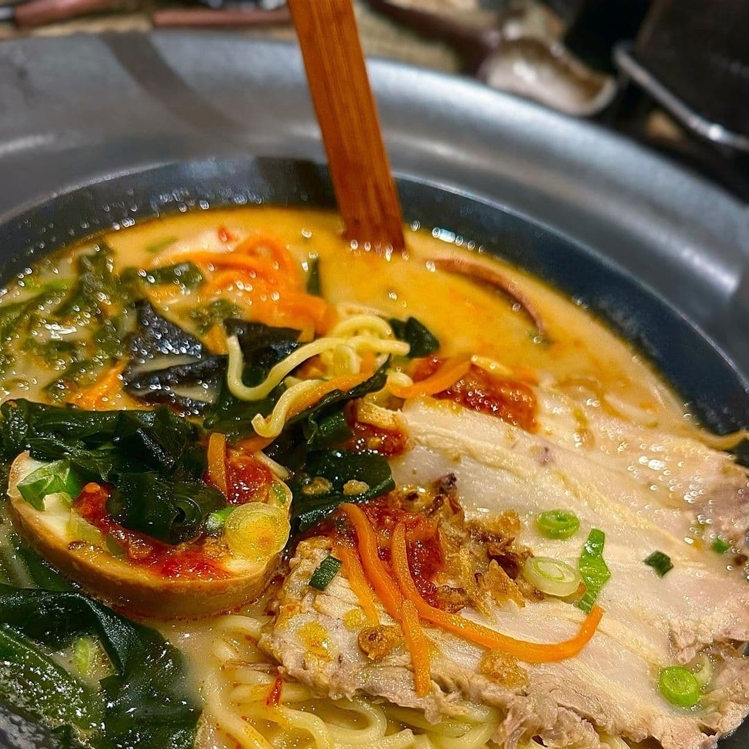 Fern Modern Sushi ramen bowl vibrant flavors delicious ingredients A close-up of a delicious bowl of ramen from Fern Modern Sushi, featuring noodles, tender slices of pork, vibrant greens, and colorful vegetables in a rich broth. A wooden spoon is seen stirring the dish, highlighting the fresh ingredients and inviting presentation.