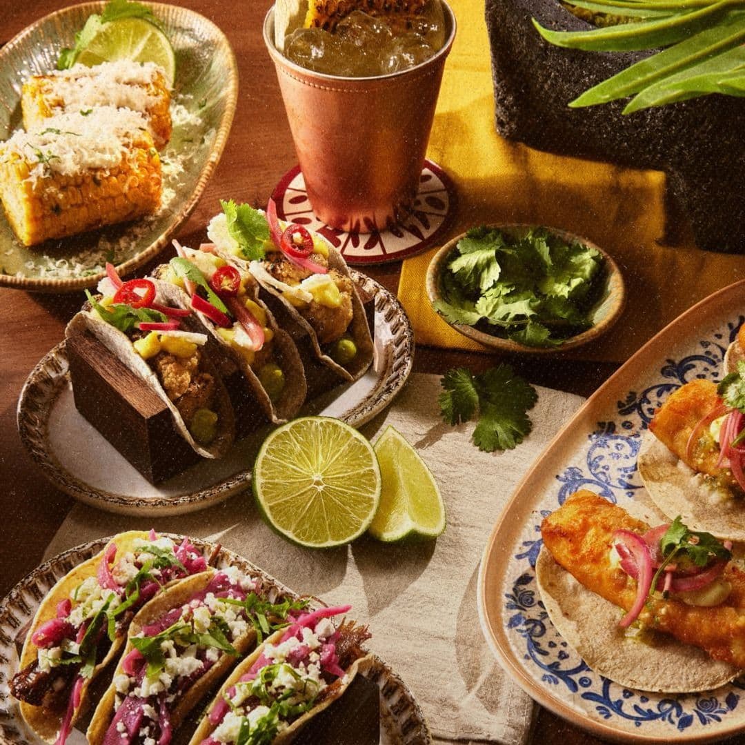 Azotea authentic mexican tacos delicious food tourism Delicious spread of tacos and Mexican street food at Azotea. The image features three types of tacos on a plate, garnished with fresh herbs and sliced chili. There are also two elote (corn on the cob) dishes, a refreshing drink in a copper cup, and lime wedges. The vibrant colors and textures highlight the rich culinary offerings of the location.