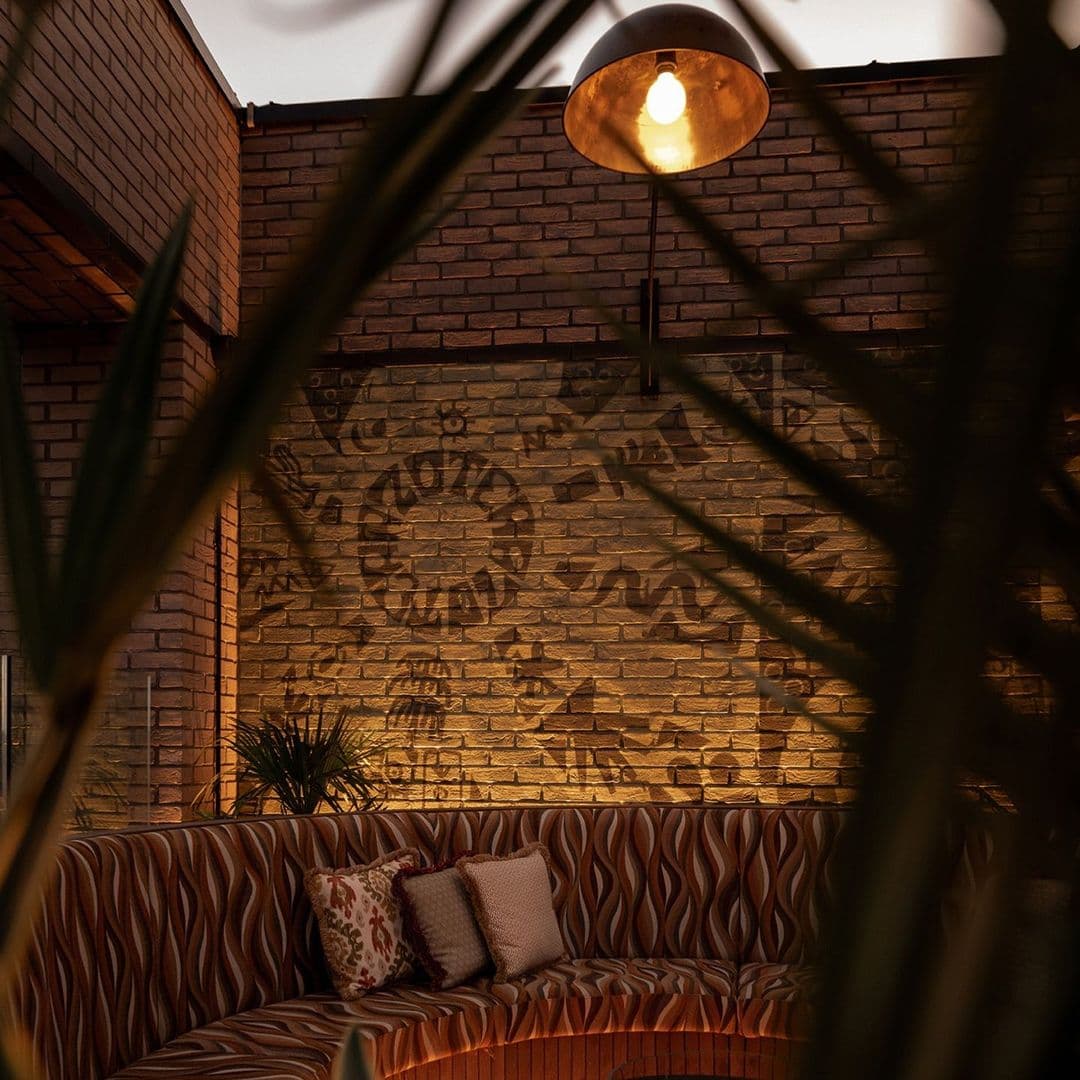 Azotea cozy interior relaxing lighting modern design Cozy seating area at Azotea, featuring a curved, patterned sofa with decorative pillows. The warm lighting highlights a textured brick wall with artistic designs, surrounded by green plants, creating an inviting atmosphere.