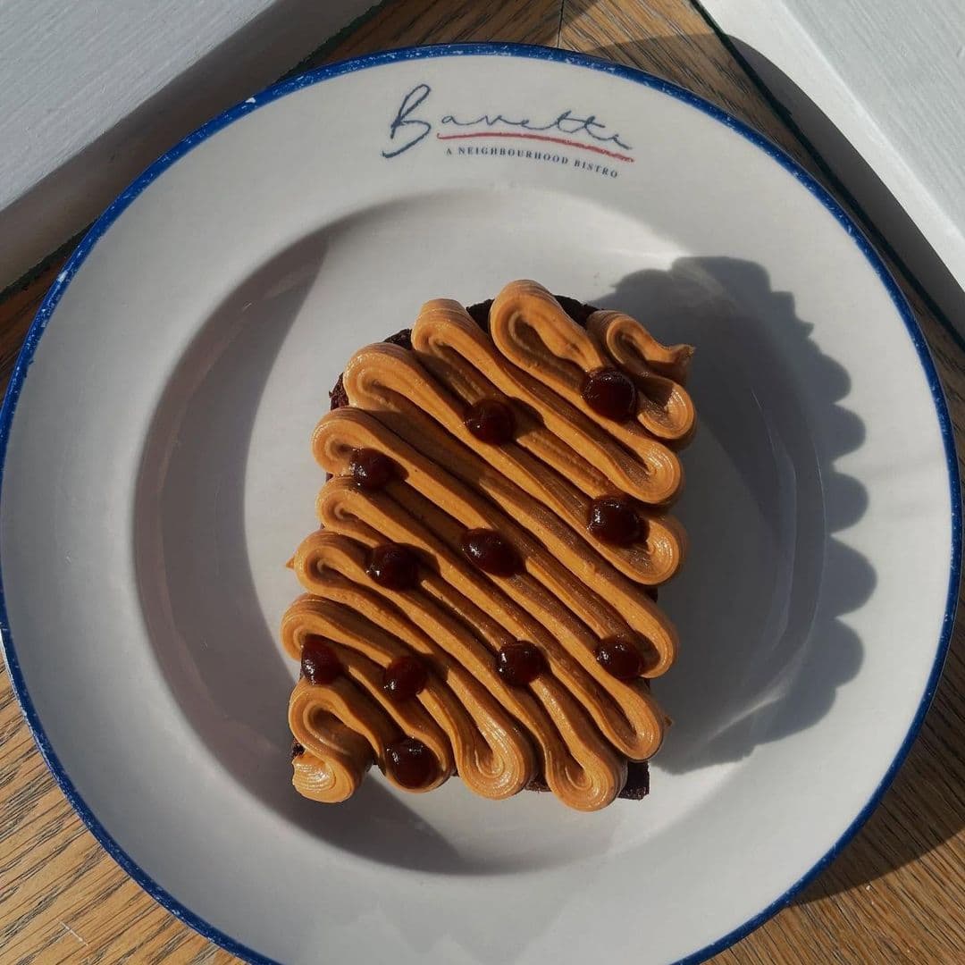 Bavette Bistro delicious dessert peanut butter cake plated Delicious dessert served at Bavette Bistro, featuring a rich cake topped with smooth, swirled caramel frosting and glossy chocolate drops, presented on a white plate with the bistro's logo.