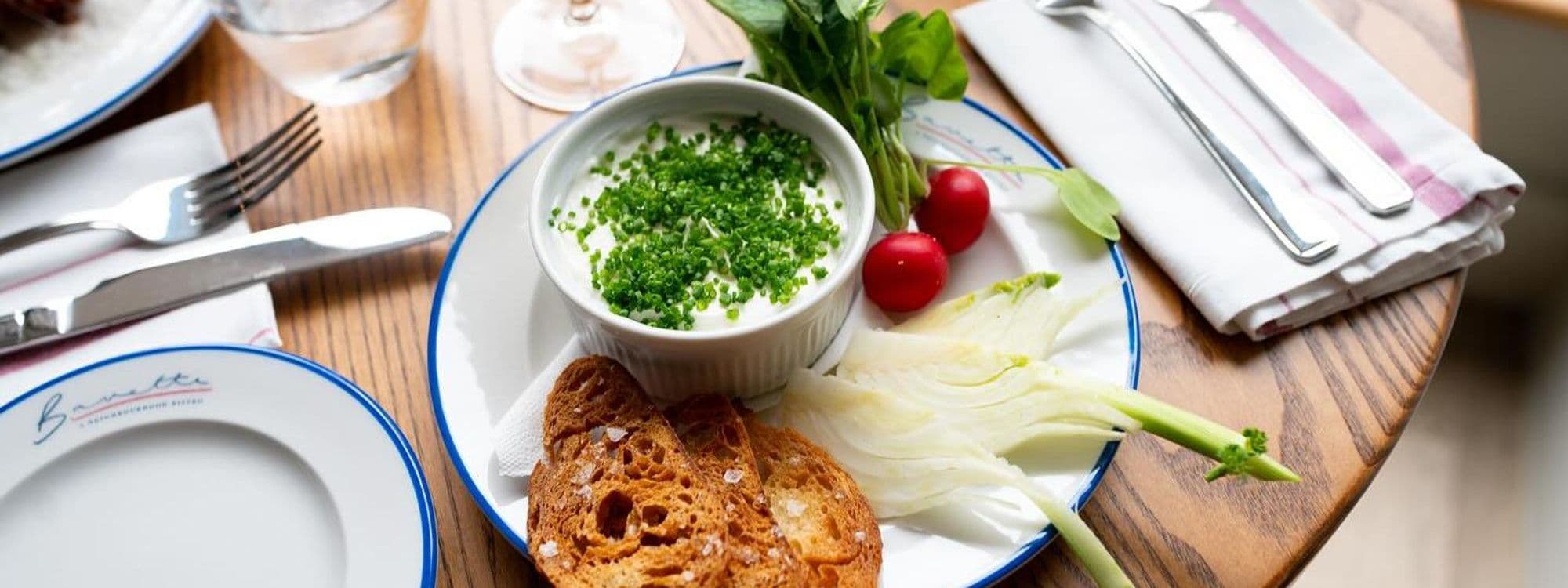 A beautifully arranged plate at Bavette Bistro featuring a bowl of creamy dip topped with fresh herbs, accompanied by crispy toasted bread, vibrant cherry tomatoes, and fresh greens. The setting includes a wooden table with elegant cutlery and a napkin, creating a warm and inviting dining atmosphere.