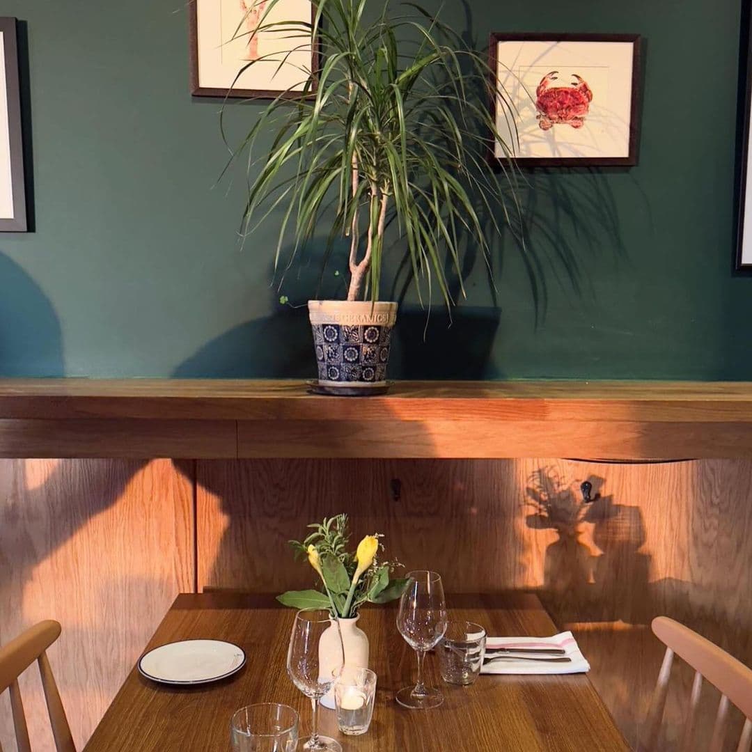 BavetteBistro cozy dining table green wall decorative plant A cozy dining setup at Bavette Bistro featuring a wooden table with a small vase of flowers, glassware, and a plate. A green wall adorned with framed artwork and a potted plant adds to the inviting atmosphere. Soft lighting creates a warm ambiance.