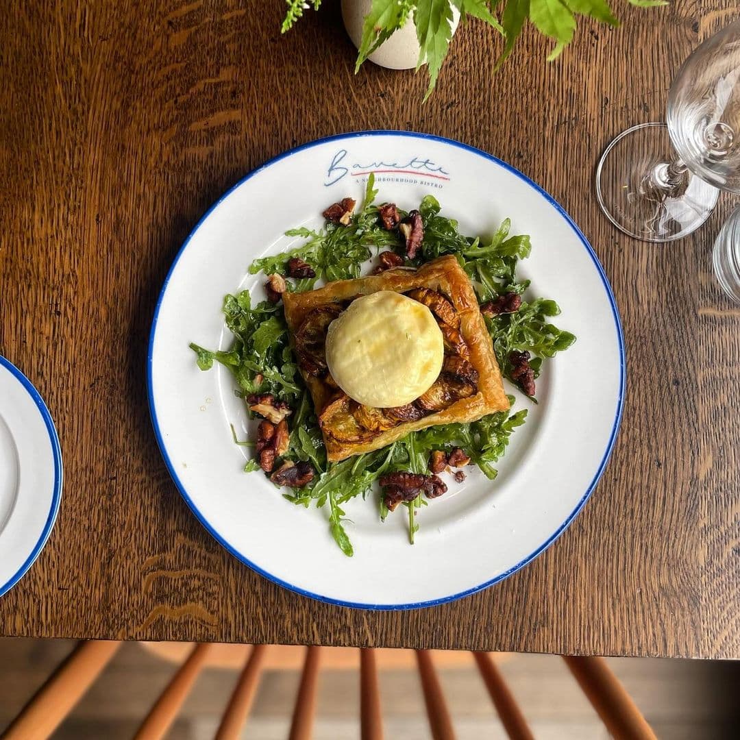 BavetteBistro gourmet dish salad arugula presentation A delicious dish served at Bavette Bistro featuring a flaky pastry topped with a creamy round element, placed on a bed of fresh arugula and sprinkled with pecans. The plate is elegantly presented on a wooden table, enhancing the dining experience.