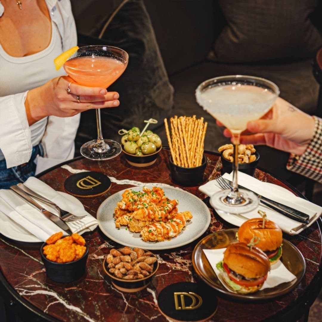Dakota Grill cocktails dining experience food platter Alt text: A table at Dakota Grill featuring a variety of dishes and drinks. Two cocktails are being raised in a toast, surrounded by plates of crispy chicken, sliders, olives, pretzel sticks, and nuts. Elegant table settings complete the inviting atmosphere.