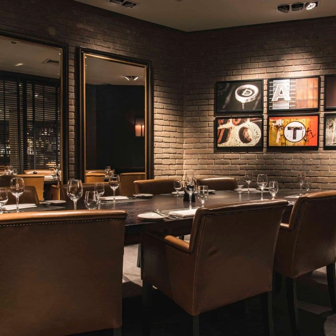 Dakota Grill elegant dining interior modern decor intimate setting Alt text: Interior of Dakota Grill featuring a stylish dining area with a large wooden table surrounded by brown leather chairs. The walls are made of exposed brick and decorated with framed artwork. Soft lighting creates a warm and inviting atmosphere.