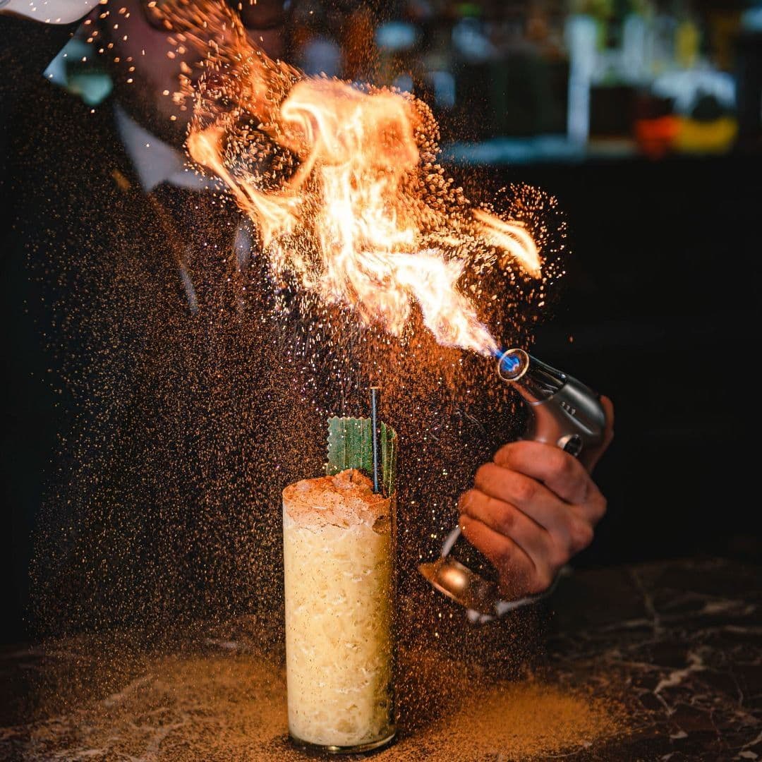 Dakota Grill flaming cocktail bartender experience drink A bartender uses a torch to ignite a cocktail at Dakota Grill, creating a dramatic flame above the drink. The cocktail is garnished with green elements and has a textured rim, surrounded by a sparkling mist.