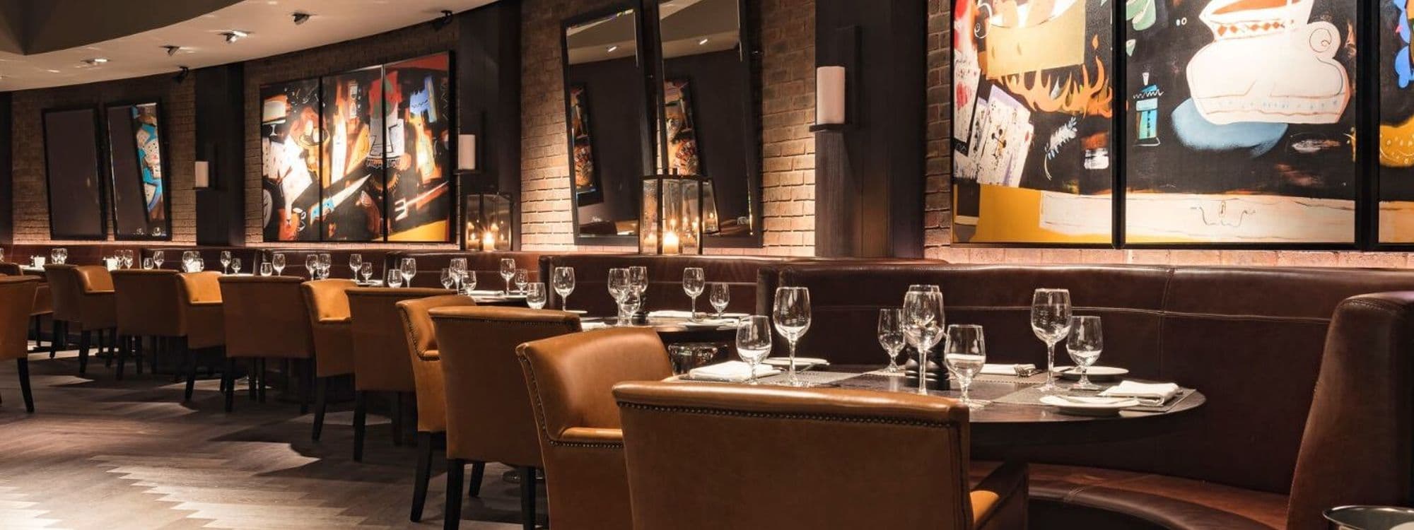 Interior of Dakota Grill featuring elegant seating with brown leather chairs and wooden tables. The walls are adorned with colorful artwork and soft lighting creates a warm ambiance. Set tables with glassware and cutlery are visible, inviting guests to enjoy a meal.