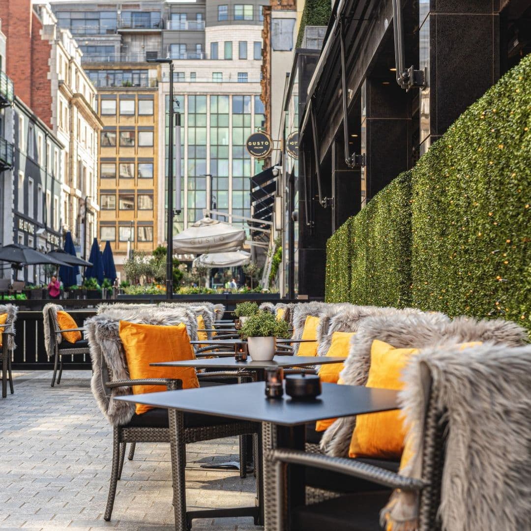 Dakota Grill outdoor dining cozy seating modern ambiance Outdoor seating area at Dakota Grill featuring stylish chairs with orange cushions and cozy fur throws. The patio is surrounded by greenery and offers a view of modern buildings in the background. Perfect for enjoying a meal in a vibrant atmosphere.
