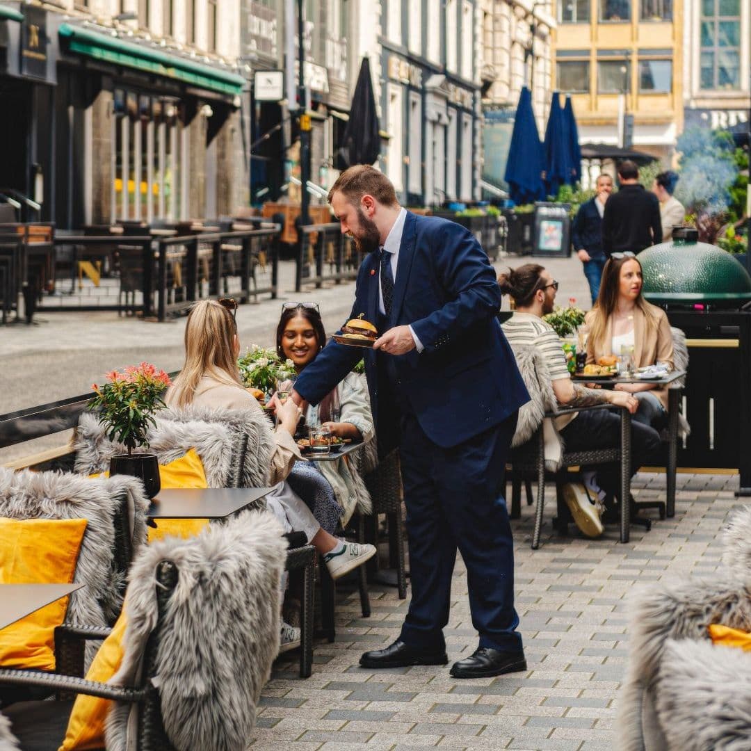 Dakota Grill outdoor dining hospitality food experience Alt text: A waiter in a suit serves food to guests at Dakota Grill's outdoor seating area. The scene features cozy chairs with fur throws, potted plants, and a lively atmosphere with people enjoying their meals and drinks.