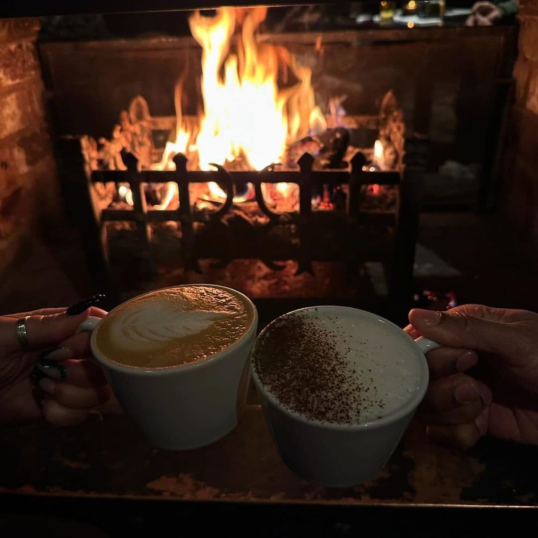 Deer Park Hotel cozy fireplace coffee cups relaxation Two hands hold steaming cups of coffee in front of a cozy fireplace, with flames dancing in the background. The setting is warm and inviting, perfect for relaxation. This image captures the comforting atmosphere at Deer Park Hotel.