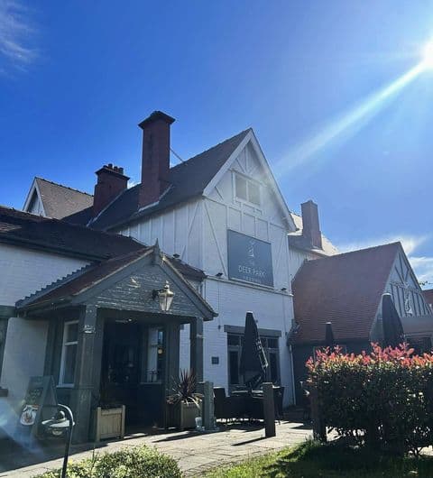 Deer Park Hotel exterior sunny day tourism attraction Alt text: The Deer Park Hotel features a charming exterior with a white facade and red accents. The building is surrounded by greenery and outdoor seating under umbrellas, with a bright blue sky above.
