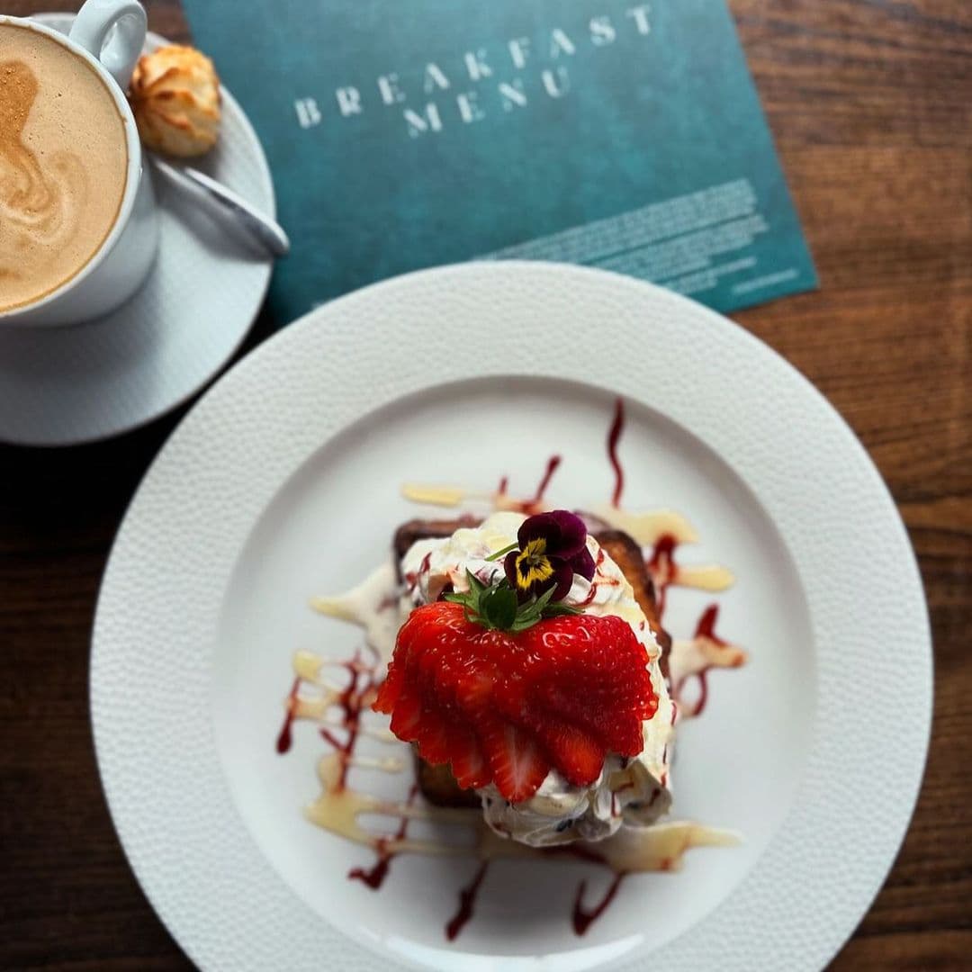 Deer Park Hotel gourmet breakfast dessert coffee strawberries Delicious breakfast dish featuring a slice of cake topped with whipped cream, fresh strawberries, and a decorative flower. A cup of coffee and a menu are visible in the background. Ideal for guests at Deer Park Hotel.