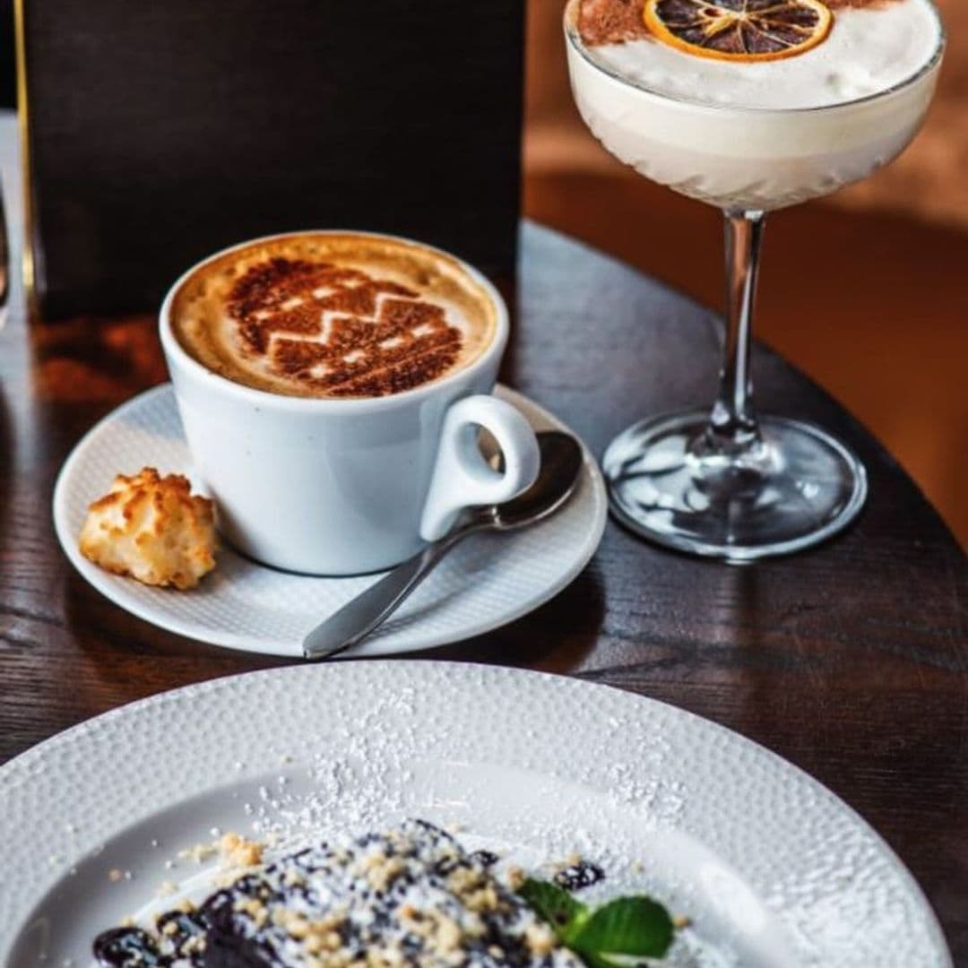 Deer Park Hotel gourmet coffee dessert luxury dining A beautifully arranged table at Deer Park Hotel featuring a cup of cappuccino with a decorative foam design, a small cookie on the side, and a dessert plate with a chocolate treat topped with powdered sugar and mint leaves. A cocktail with a lemon garnish is also visible in the background.