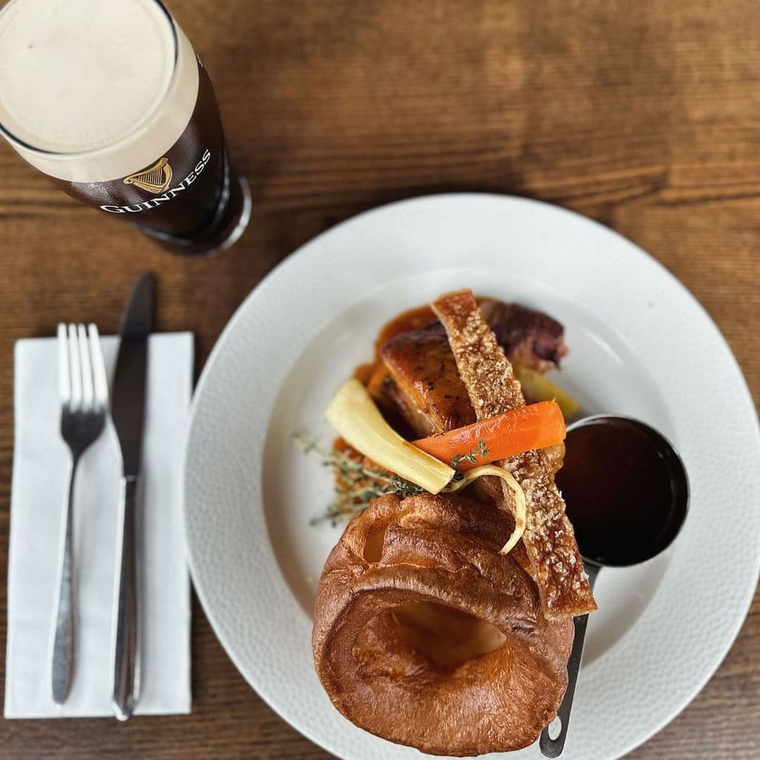Deer Park Hotel gourmet dining Guinness roast beef Yorkshire pudding A delicious plate of roast pork with crispy crackling, accompanied by carrots, parsnips, and a rich sauce. A large Yorkshire pudding sits beside the meat. A glass of Guinness beer is visible in the background. This meal is served at Deer Park Hotel.