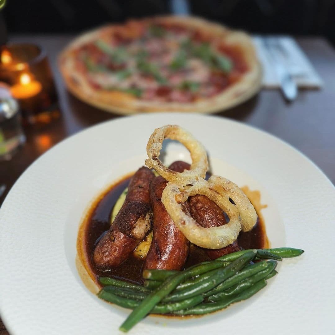 Deer Park Hotel gourmet dining sausages onion rings green beans Delicious plate of sausages served with green beans and crispy onion rings, with a pizza in the background. Enjoy a meal at Deer Park Hotel.