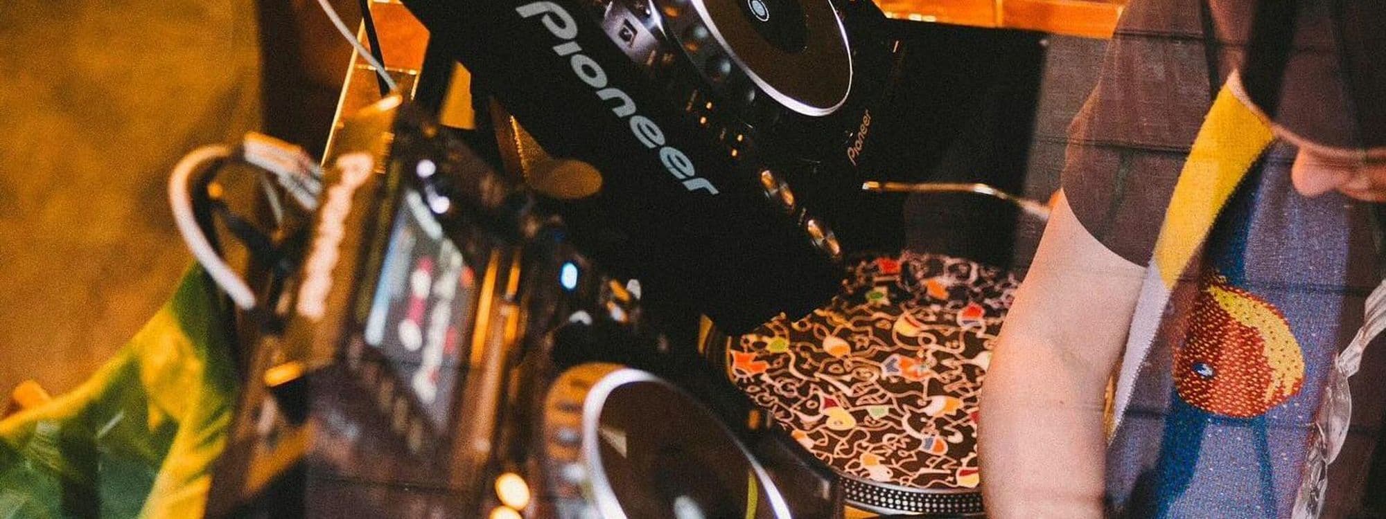 Alt text: A DJ is mixing music at a vibrant venue called Holding Patterns. The image shows DJ equipment, including turntables and a mixer, with colorful decorations in the background.